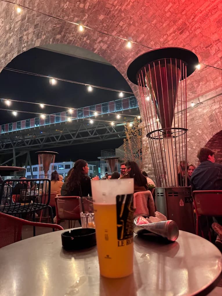 Terrasse des Arches la nuit sous les guirlandes, voûte éclairée de rouge, vue sur le pont du métro m2.
