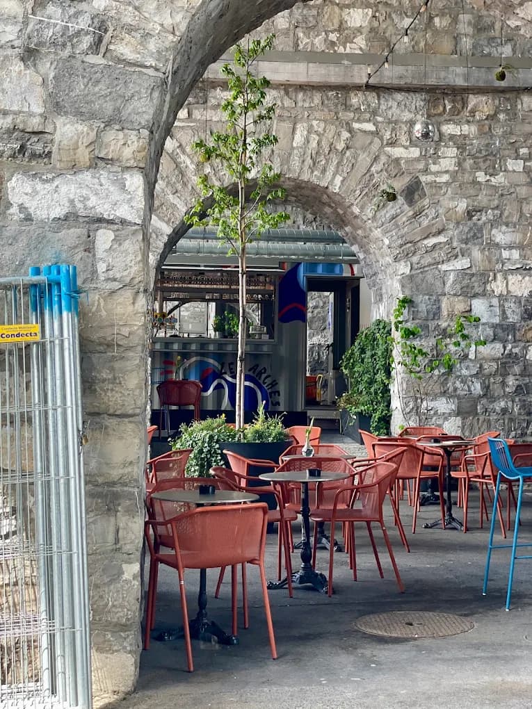 Terrasse des Arches de jour : chaises rouges et plantes sous une arche de pierre, Lausanne.